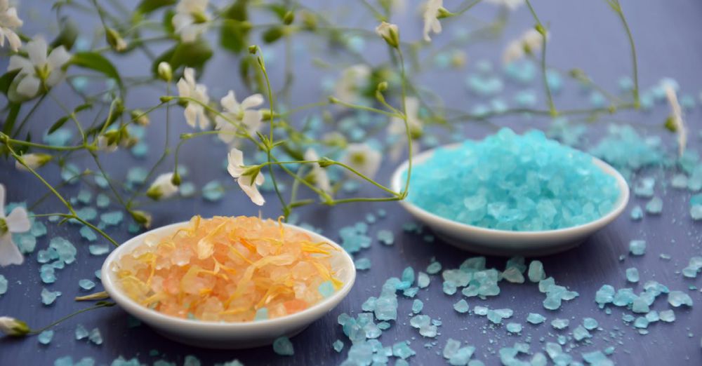 Bath Salts - A close-up of colorful bath salts in dishes surrounded by delicate white flowers on a blue surface.