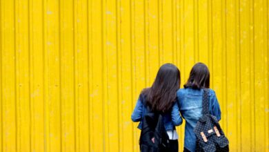 Backpacks - Two women wearing denim jackets and backpacks stand against a vibrant yellow wall.