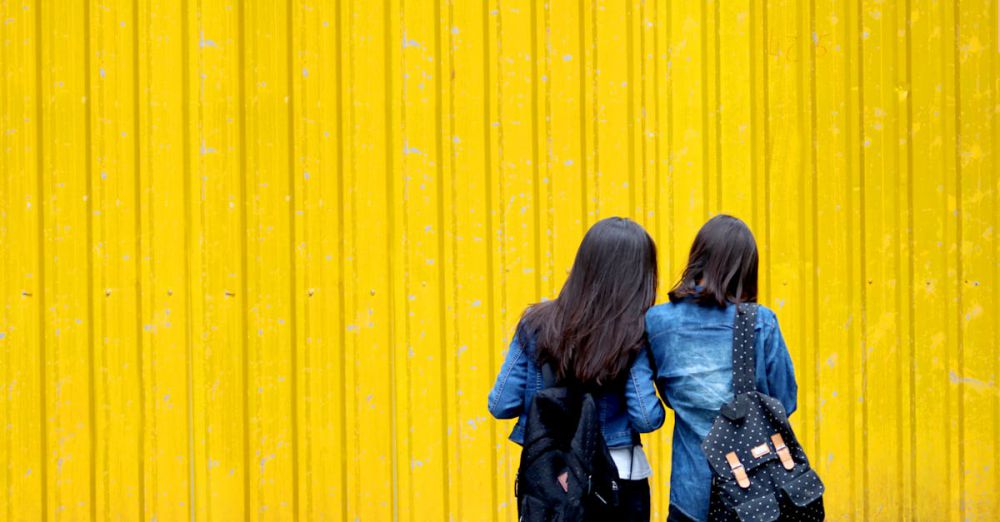 Backpacks - Two women wearing denim jackets and backpacks stand against a vibrant yellow wall.