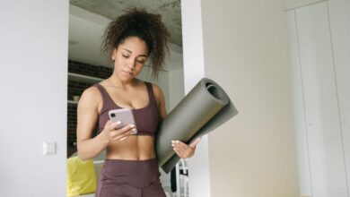 Mindfulness Apps - Young woman in activewear holding yoga mat and smartphone, preparing for a home workout.