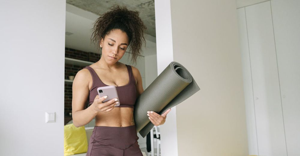 Mindfulness Apps - Young woman in activewear holding yoga mat and smartphone, preparing for a home workout.