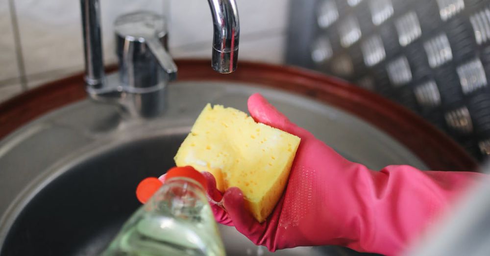 Dishwashing Liquids - Hands wearing rubber gloves pouring detergent on a sponge over a kitchen sink.