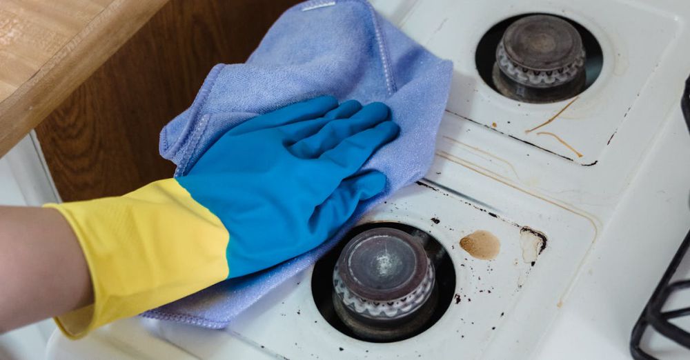 Cleaning Products - A person wearing rubber gloves cleaning a dirty gas stove with a cloth and spray bottle.