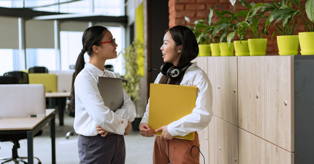 Document Holders - Two young professional women engaging in a conversation in a modern office setting.