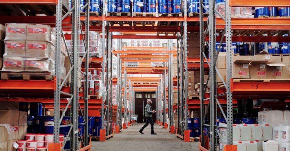 Organization Products - A man walking through a large industrial warehouse with stacked shelves filled with goods and products.