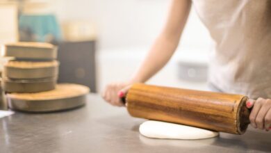Baking Pans - A woman using a wooden rolling pin to flatten dough in a bakery kitchen setting.