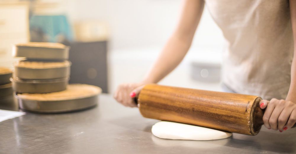 Baking Pans - A woman using a wooden rolling pin to flatten dough in a bakery kitchen setting.