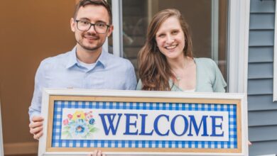 Key Holders - A cheerful couple stands at their new home entrance holding a welcome sign.