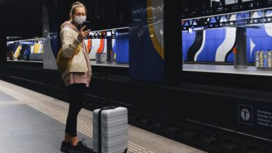 Carry-On Luggage - A traveler wearing a mask checks her phone on a subway platform during the pandemic.