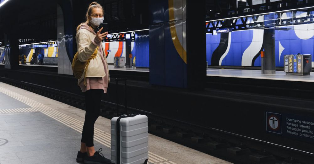 Carry-On Luggage - A traveler wearing a mask checks her phone on a subway platform during the pandemic.