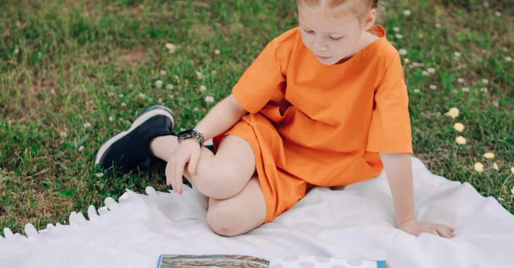 Storybooks - A child in an orange outfit reading books outdoors on a blanket. Perfect for education and leisure themes.