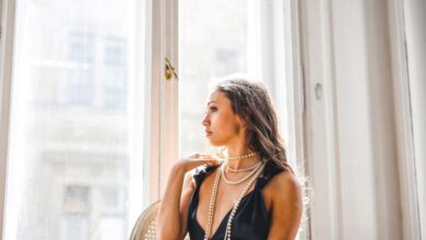 Necklaces - Stylish young woman in black dress and pearls sitting by a sunlit window indoors.