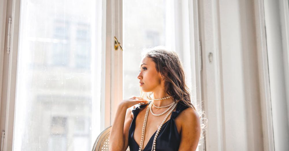 Necklaces - Stylish young woman in black dress and pearls sitting by a sunlit window indoors.