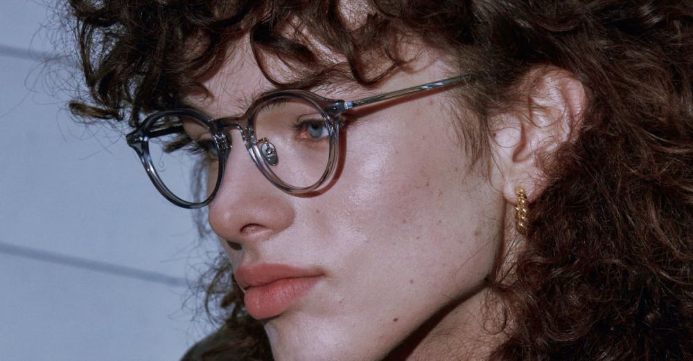 Gold-Plated Accessories - Close-up portrait of a young adult with curly hair wearing vintage glasses and earrings.